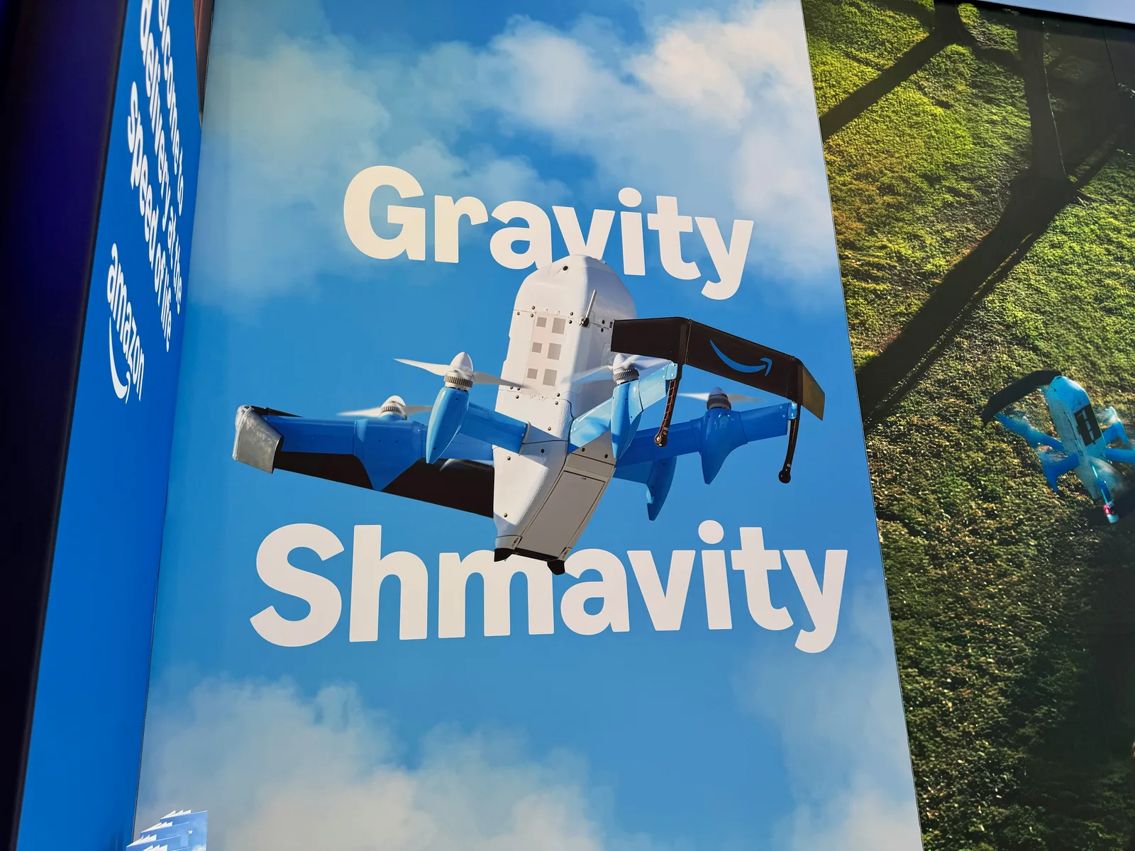 Amazon Prime Air MK30 delivery drone pictured on a large trade show banner reading Gravity Shmavity, with a second MK30 flying over green fields on the adjacent panel
