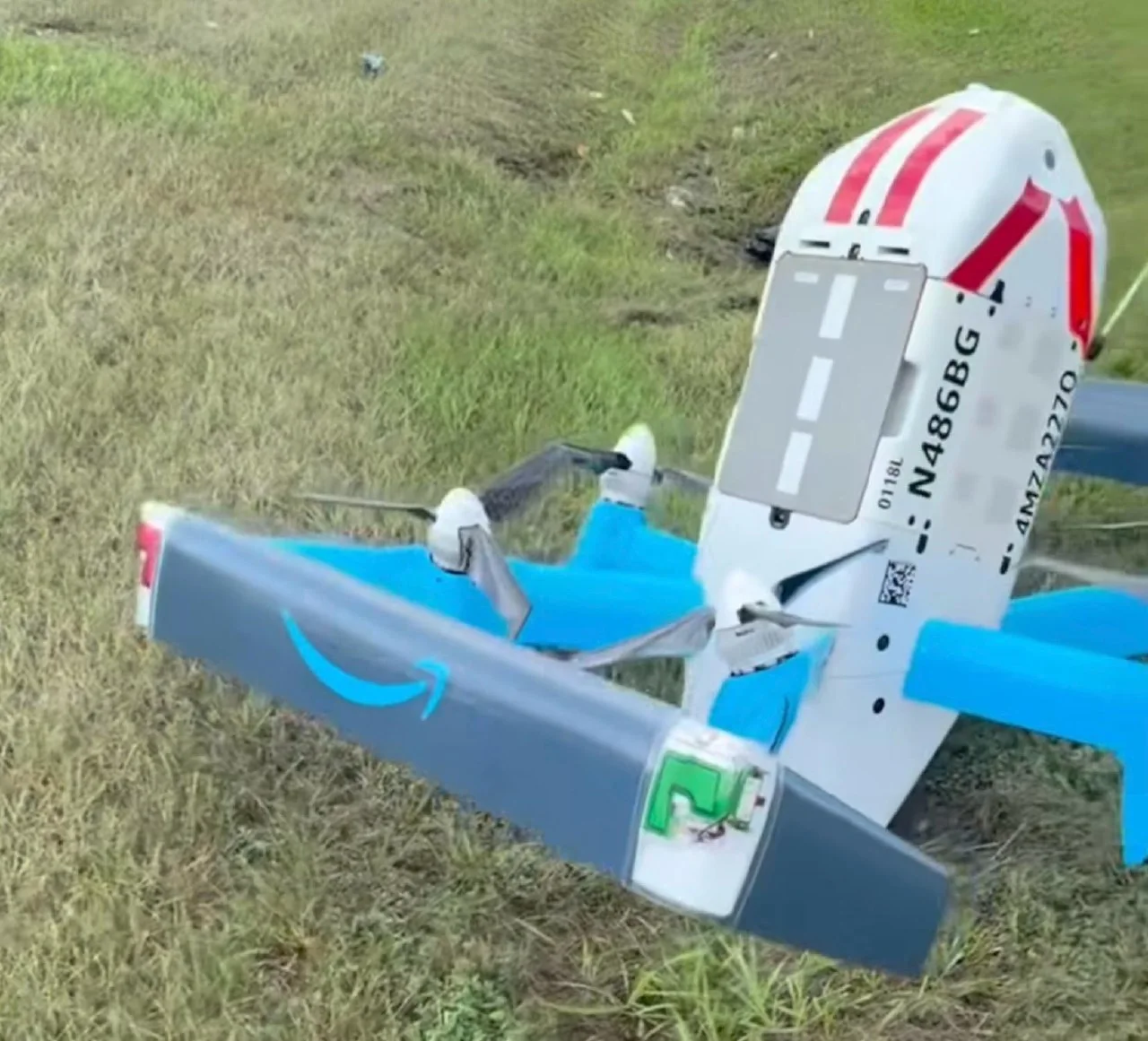 Close-up of the Amazon Prime Air MK30 drone N486BG resting on its wingtip in the grass, Amazon smile logo and a green light module visible on the wing