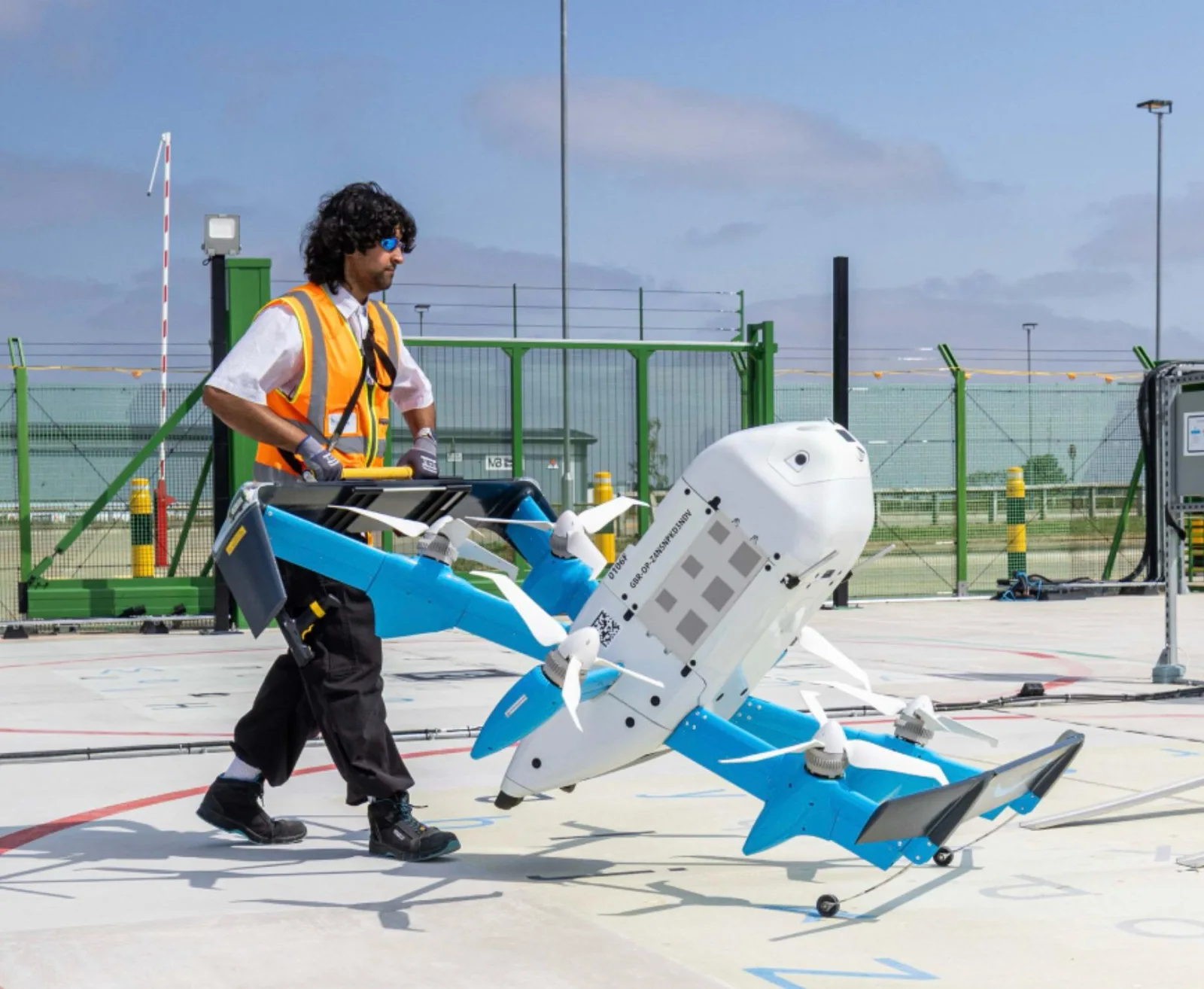 A ground handler in an orange high-visibility vest wheels an Amazon Prime Air MK30 delivery drone across a painted launch pad using a yellow handling bar