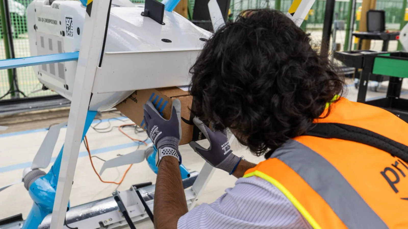 An Amazon worker in a high-visibility vest loads a cardboard package into the underside of a Prime Air delivery drone