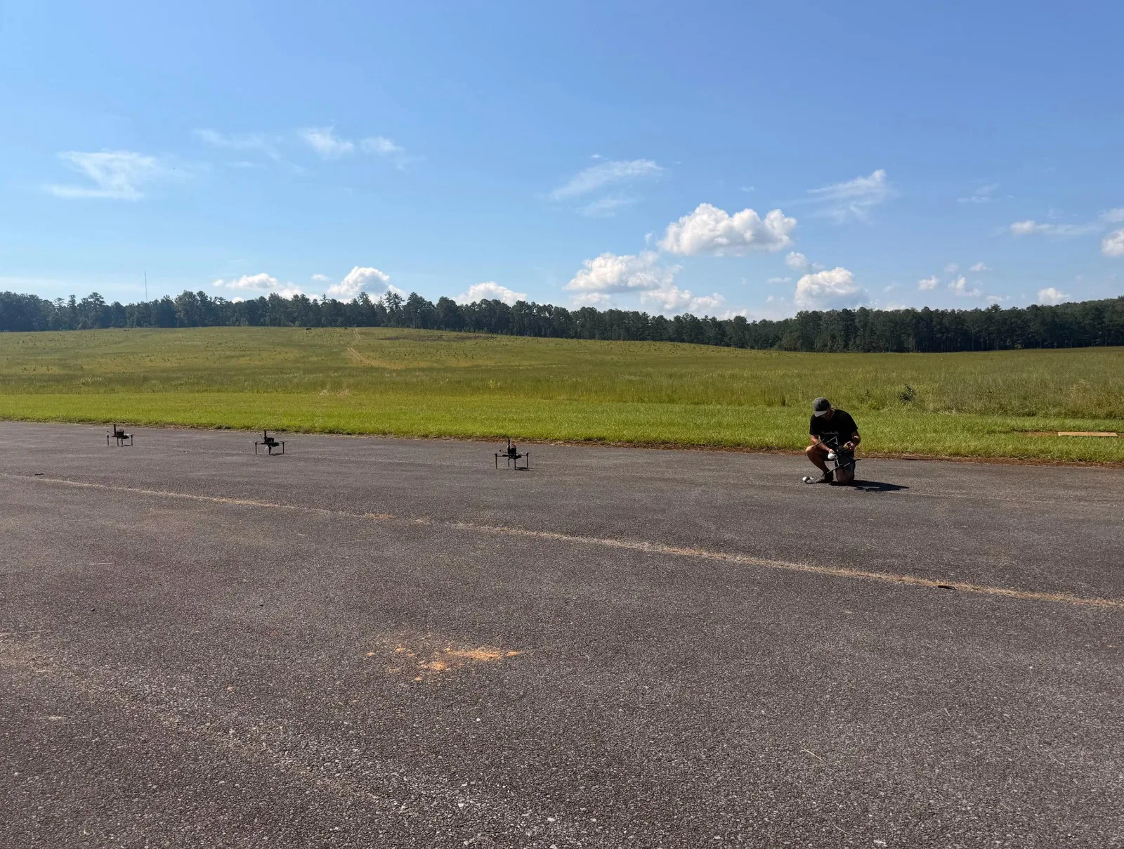 Three quadcopters on an asphalt strip beside a grass field, with a team member kneeling to prepare a fourth drone