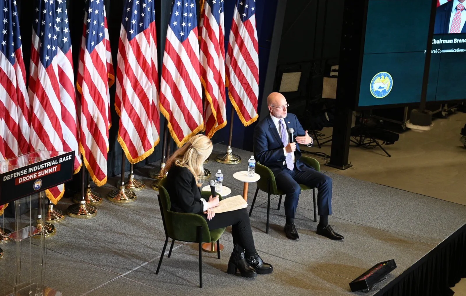 Brendan Carr speaking on stage at the Defense Industrial Base Drone Summit, overhead shot with American flags and the Small Business Administration seal on screen