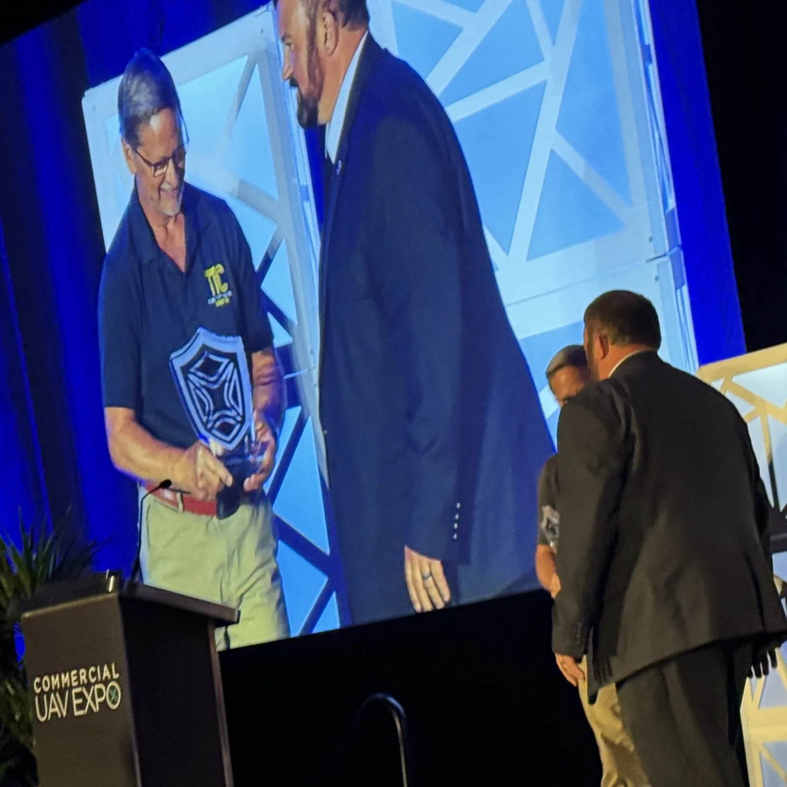 Projected on a large screen, Greg Whiting in a navy polo shirt with a yellow TIC logo accepts a shield-shaped glass award and shakes hands with Matt Rowland in a dark suit