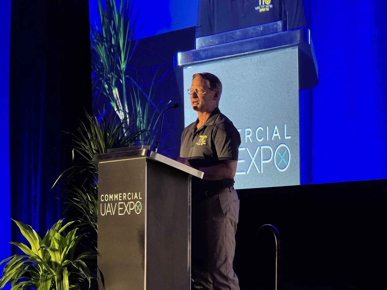 Greg Whiting speaks from the Commercial UAV Expo lectern wearing a dark polo with a yellow Technology Integration Cell logo, his name projected on the screen behind him