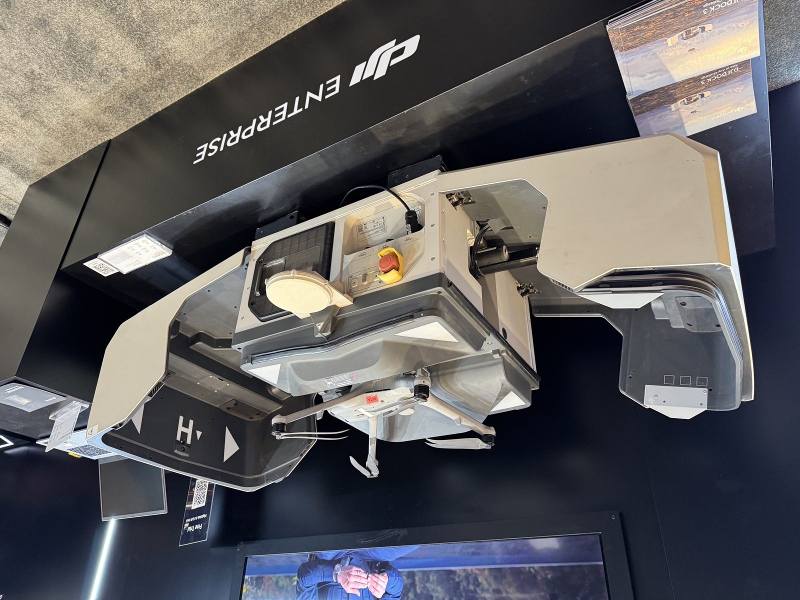 Cutaway DJI Dock 3 on the DJI Enterprise stand at Commercial UAV Expo 2026 with a Matrice 4D series aircraft loaded on the landing pad, Dock 3 brochures and a laptop in frame