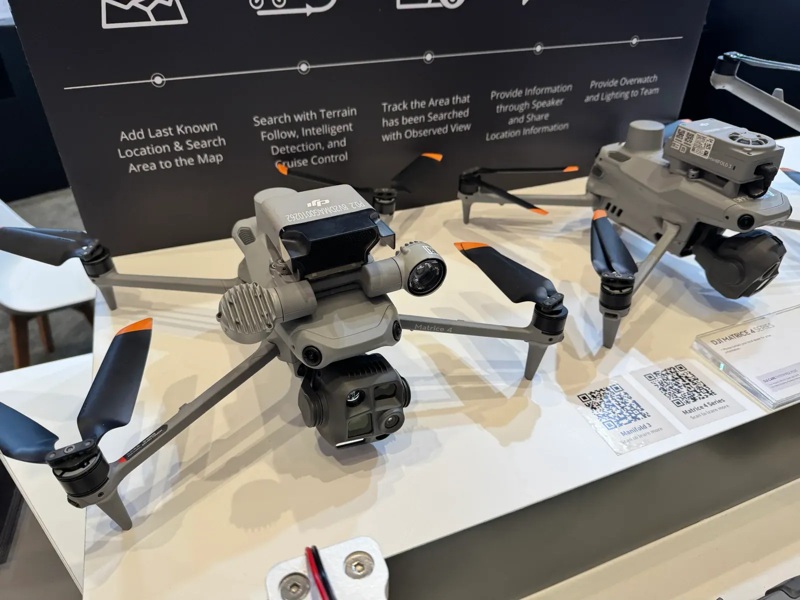 DJI Matrice 4 Series drone with gimbal camera and Manifold 3 computer on display at the DJI Enterprise booth, in front of a panel listing search-and-rescue features
