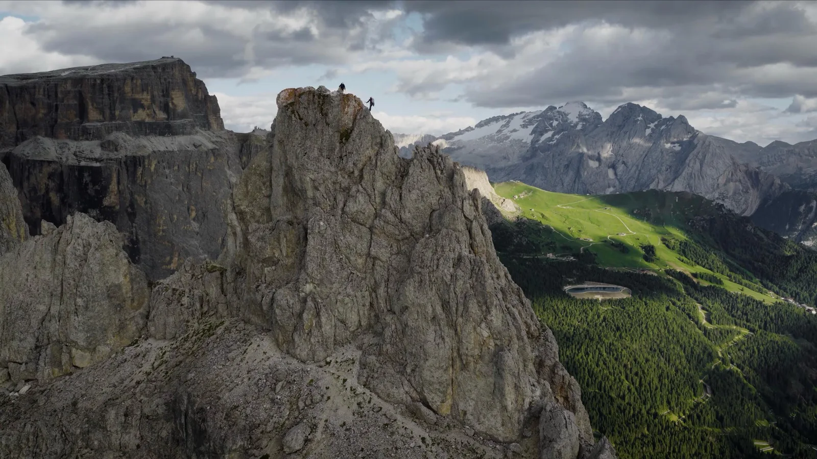 Two climbers stand on top of a jagged rock spire in the Dolomites, seen from a drone