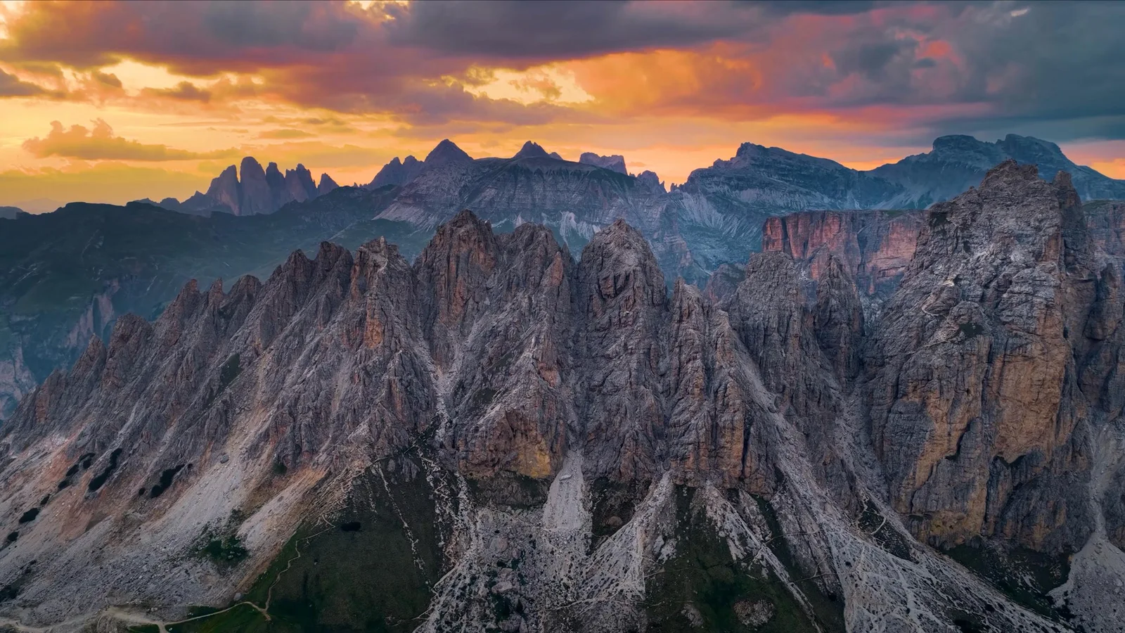 Jagged Dolomites peaks under an orange and purple sunset sky, aerial view