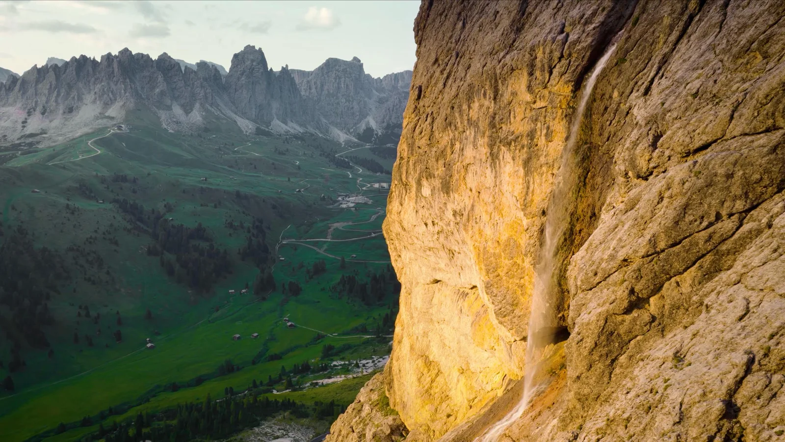 Aerial view of a waterfall running down a sunlit limestone cliff beside a green Dolomites valley