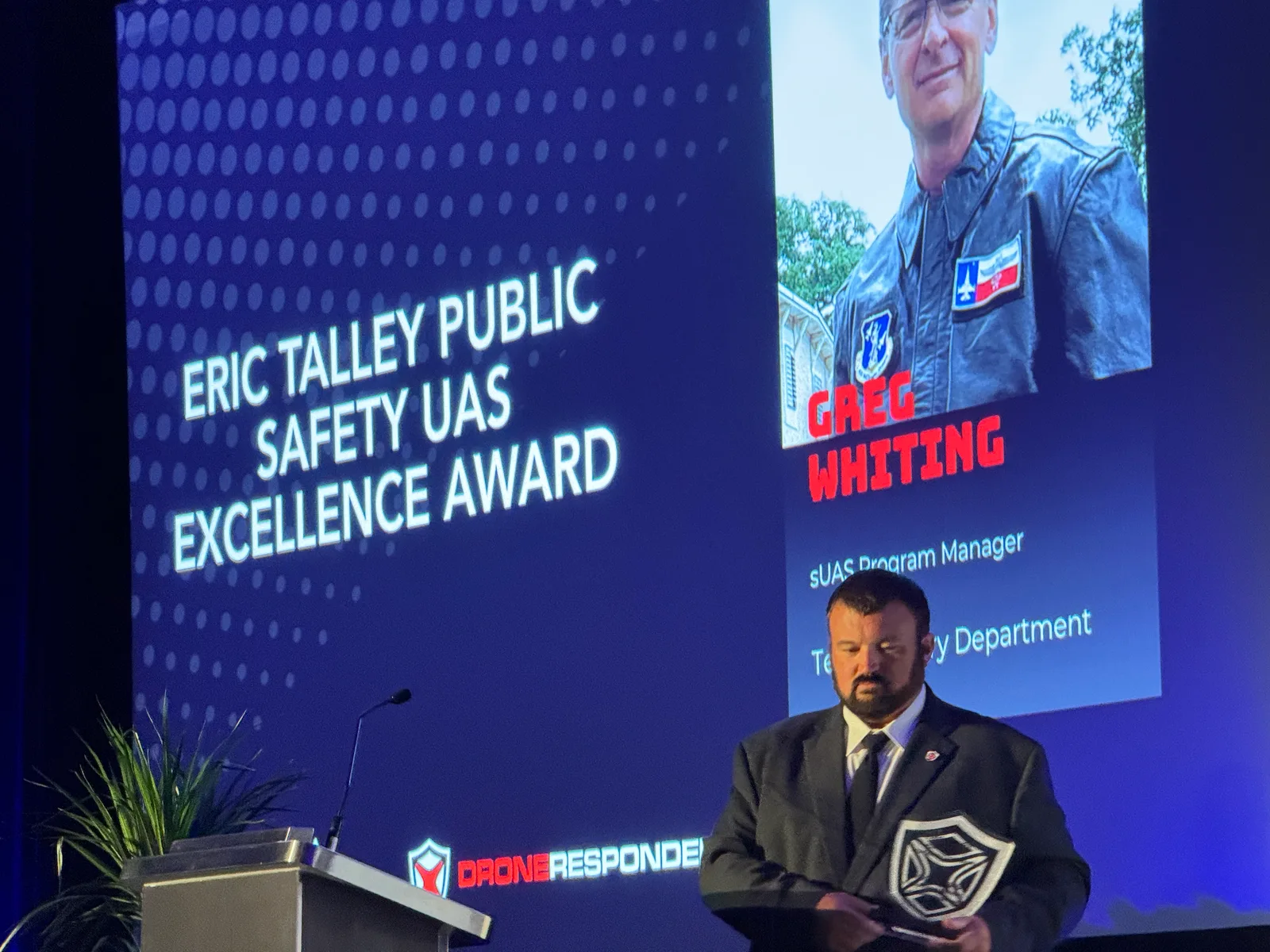 Matt Rowland in a dark suit stands at a lectern holding a shield-shaped award while a screen behind him reads Eric Talley Public Safety UAS Excellence Award above a photo of Greg Whiting
