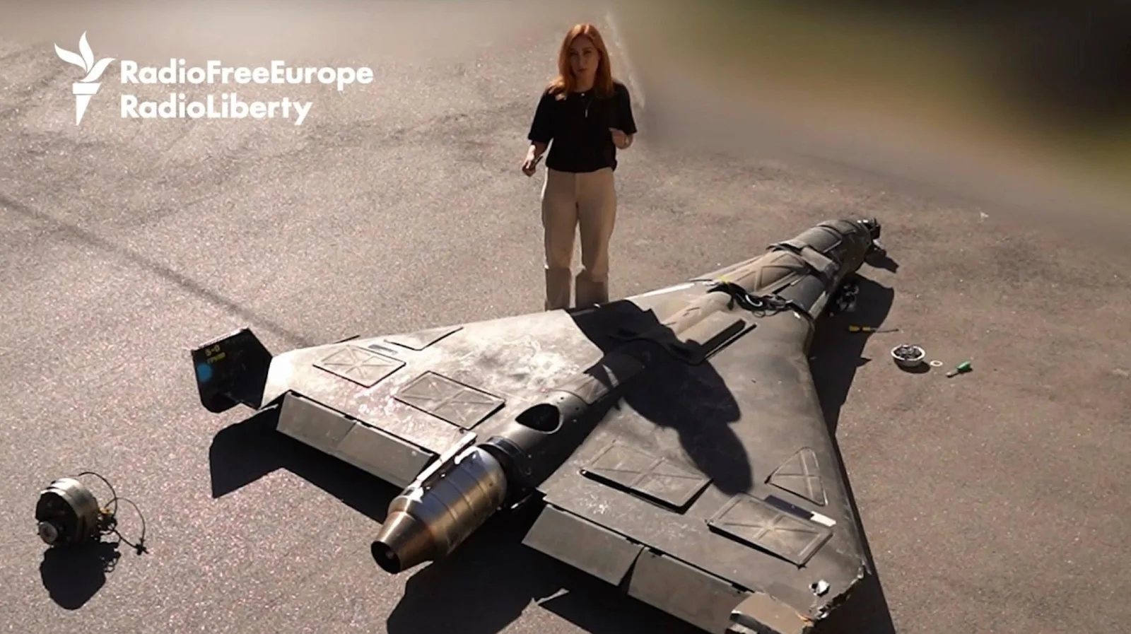 Overhead view of a dark delta-wing Russian Geran-4 Seeker attack drone lying on asphalt with its turbojet engine at the rear, a journalist standing beside it and a removed component on the ground, from RFE/RL's September 2026 teardown video