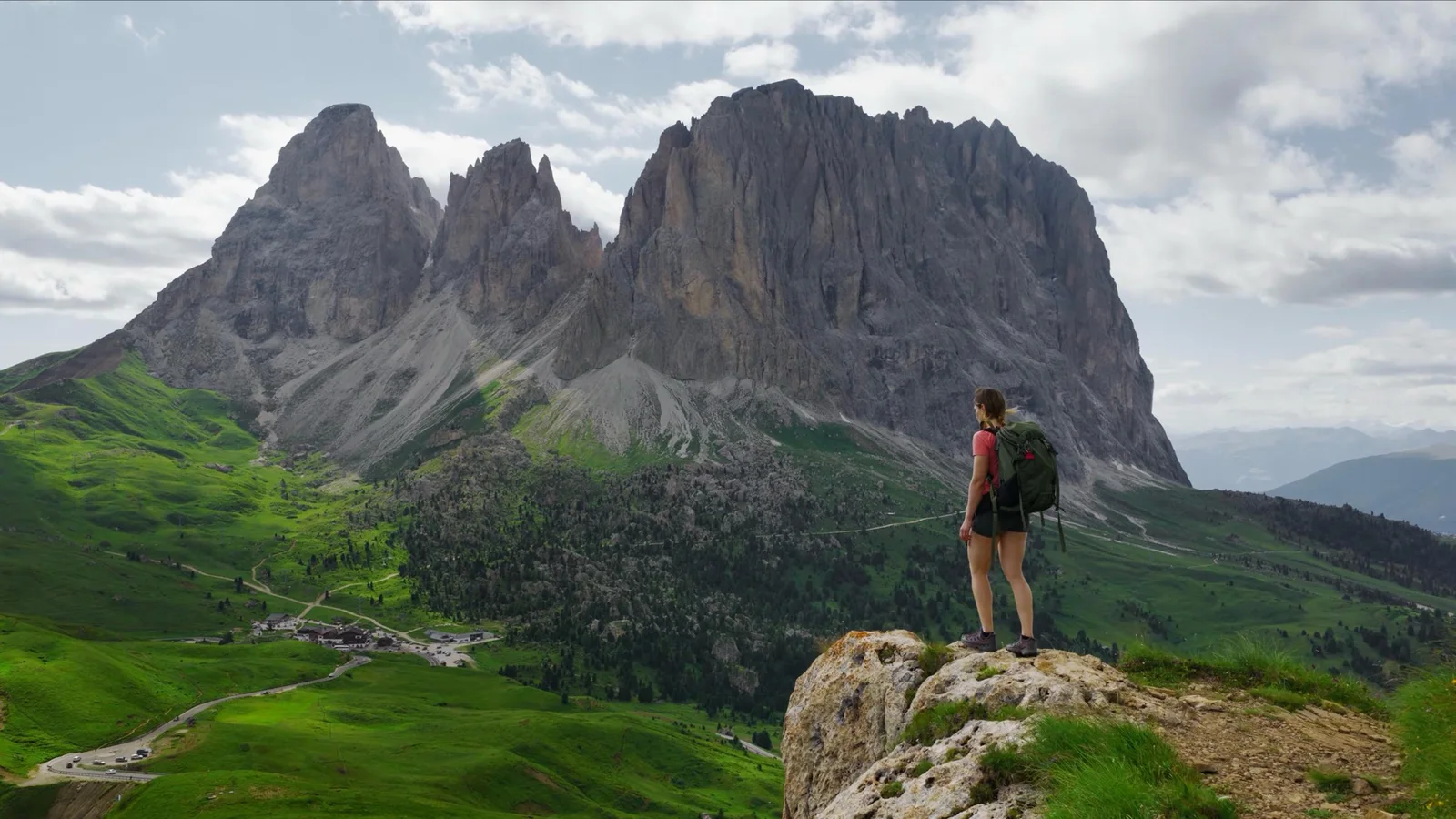 A hiker with a green backpack stands on a rocky outcrop facing a massive Dolomites rock wall above green alpine meadows
