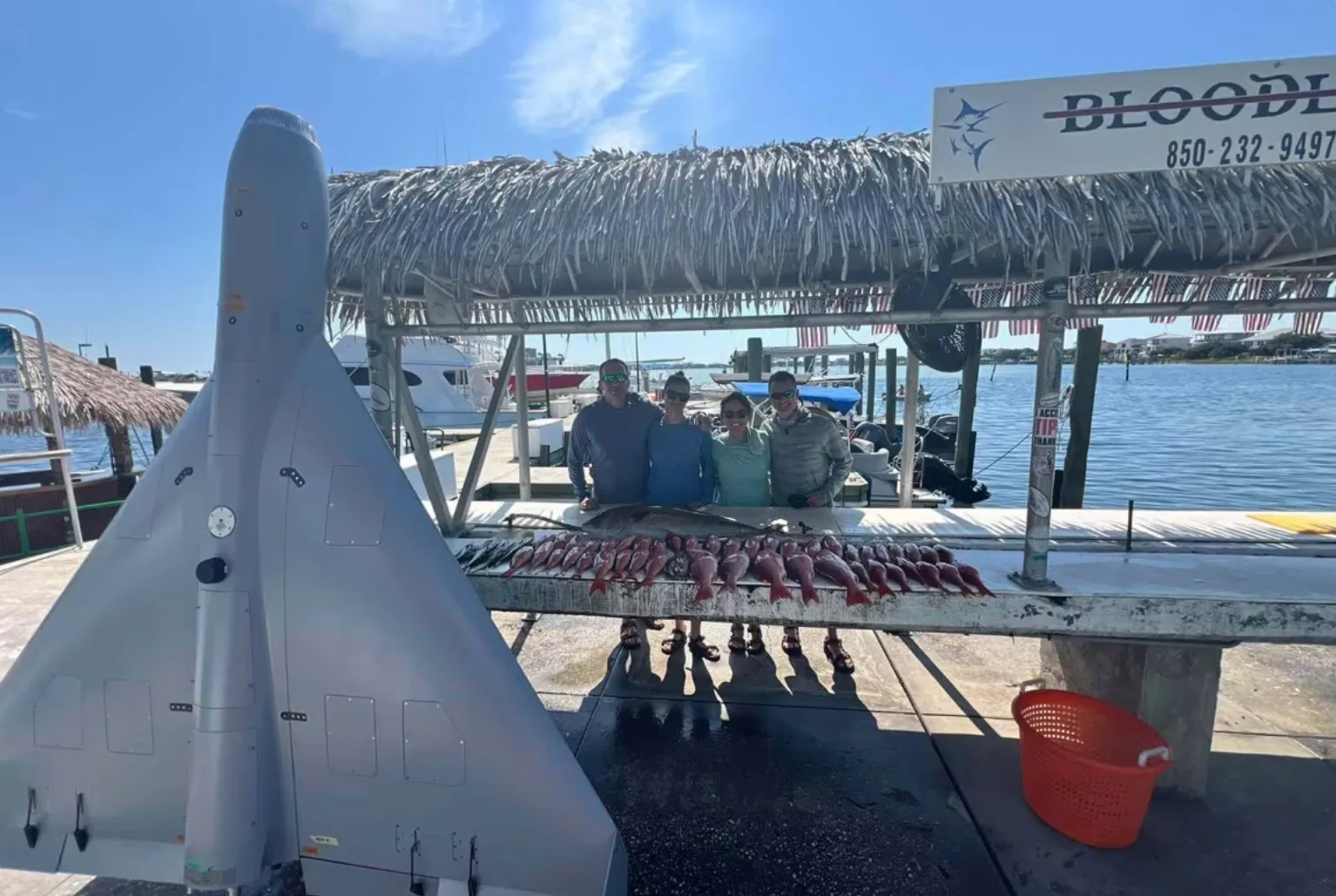 Gray delta-wing drone standing on a dock beside a fish-cleaning table lined with red fish, four anglers posing behind it under a thatched roof