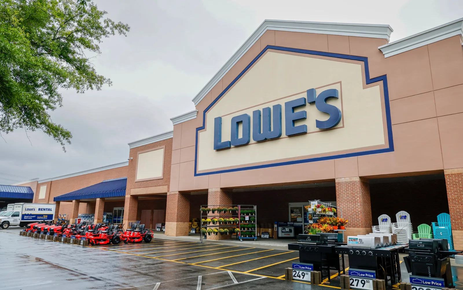Front of a Lowe's store with the blue Lowe's sign, red riding mowers and grills lined up outside, and a Lowe's rental truck parked at the left