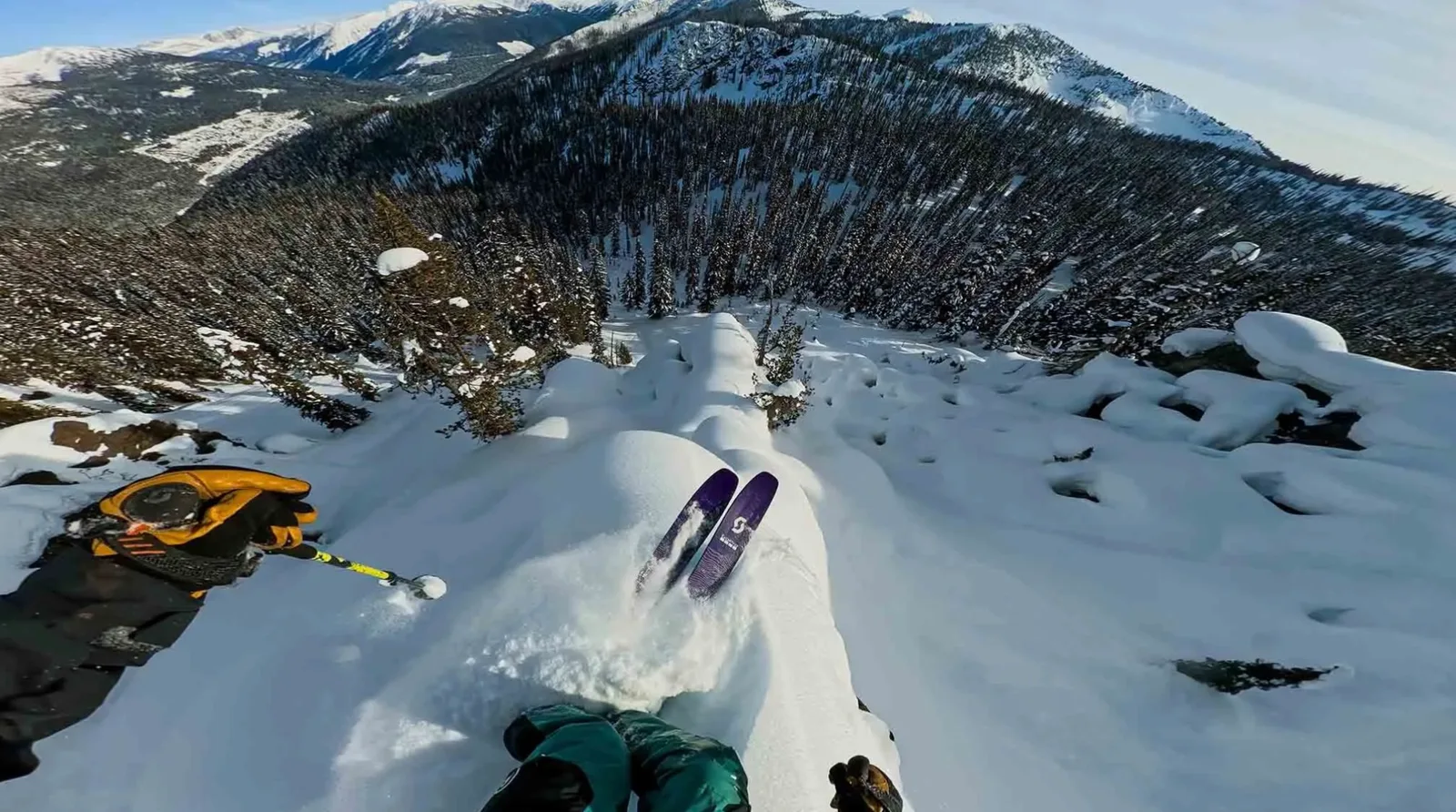 First-person GoPro view of a skier dropping into a deep powder line above a forested valley, with snow-covered peaks on the horizon.