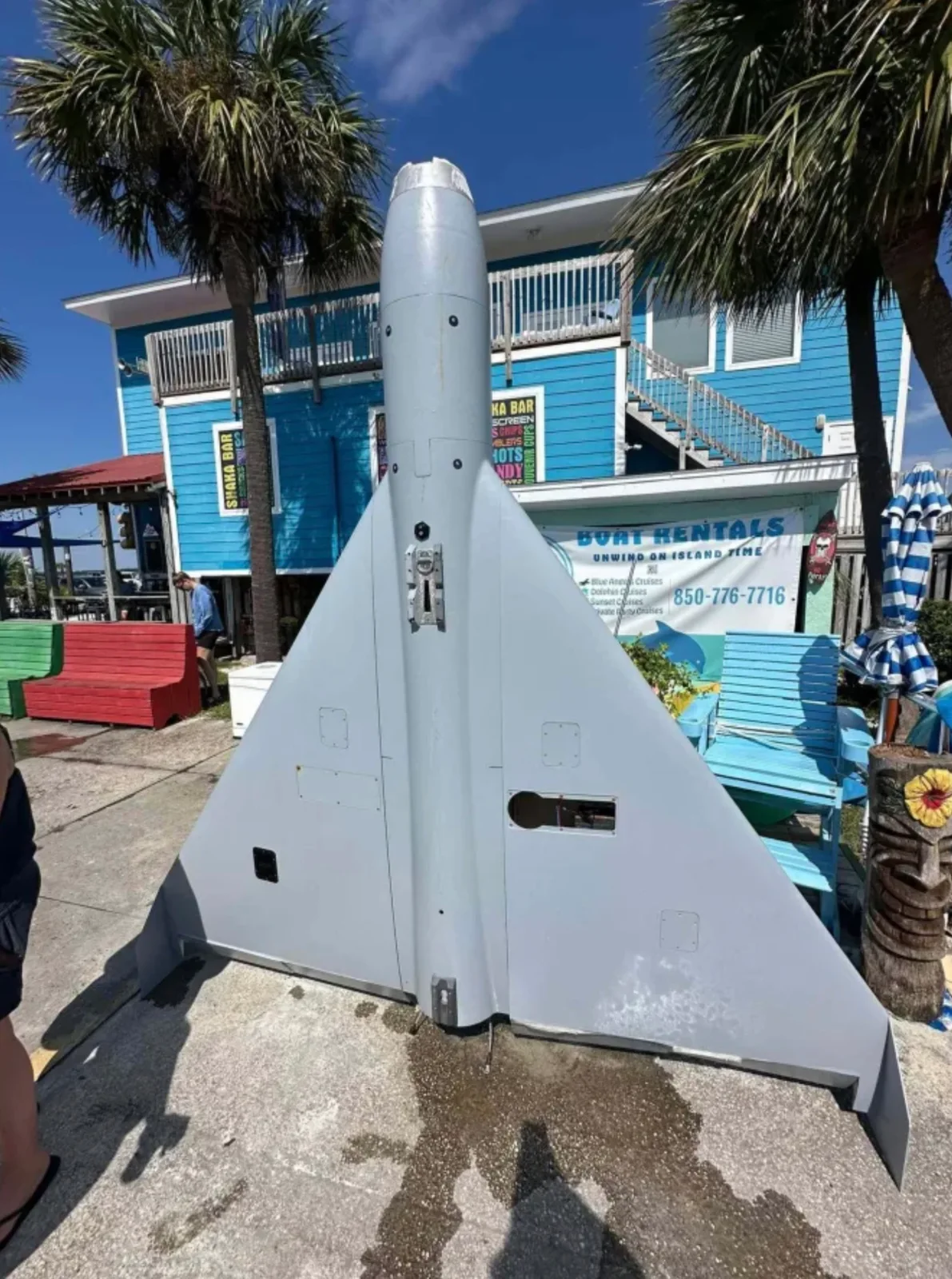 Gray delta-wing drone with a missing nose cone standing upright on a dock in front of a blue two-story bar and a Boat Rentals sign