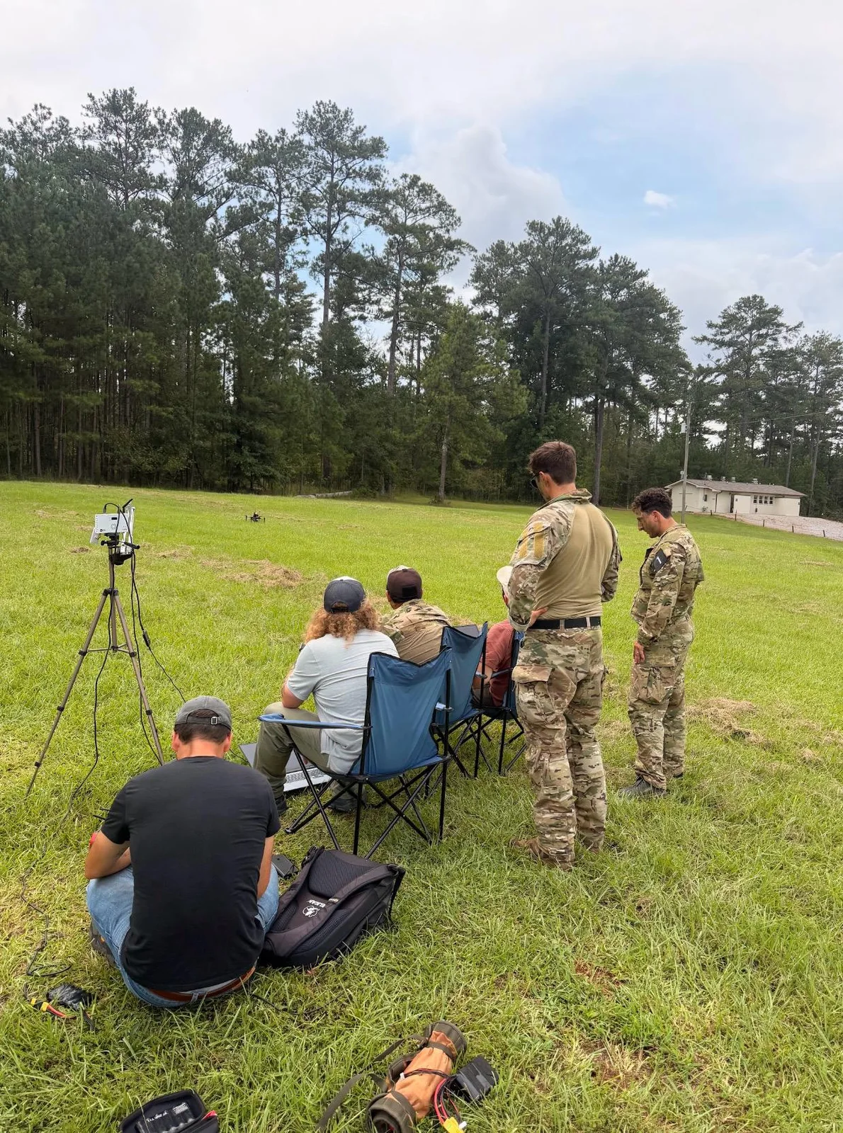 Two soldiers in camouflage uniforms stand behind seated operators with a laptop while a quadcopter hovers low over a grass field, with a tripod-mounted ground unit on the left