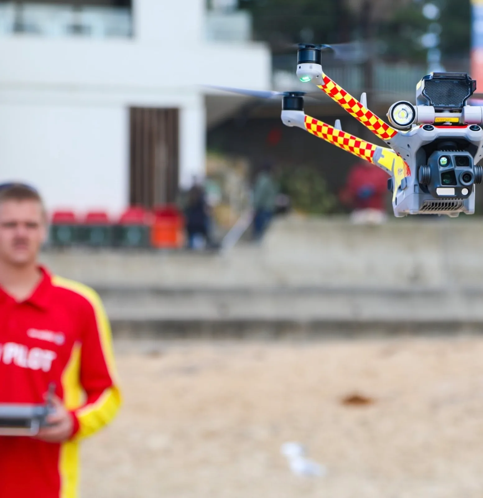 A lifesaving drone fitted with a spotlight and loudspeaker hovers above a beach while a lifeguard in a red and yellow pilot shirt flies it with a handheld controller