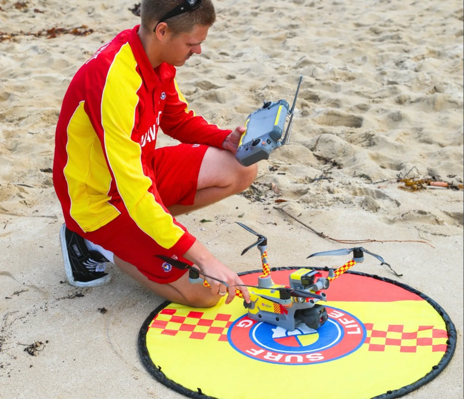 Surf Life Saving NSW lifeguard in red and yellow uniform kneeling on beach sand, holding a controller beside a red-and-yellow DJI drone on a landing pad