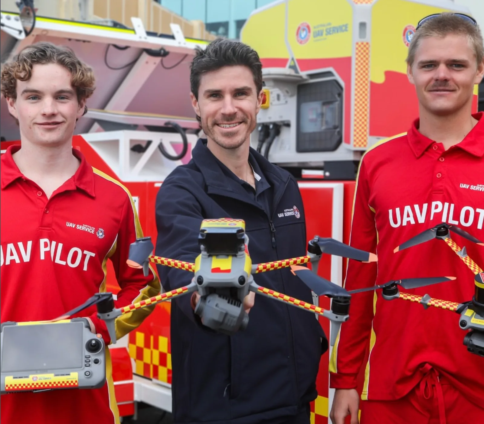 Three Surf Life Saving NSW UAV Service pilots in red and navy uniforms hold a yellow and red DJI Matrice-style quadcopter and controller in front of a marked response vehicle