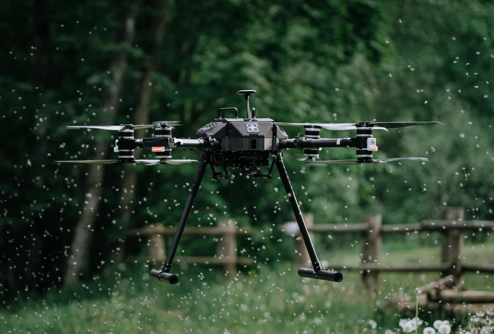 Black XTEND quadcopter with carbon-fiber landing legs and a gimbal camera hovering low over a meadow as dandelion seeds swirl through the air