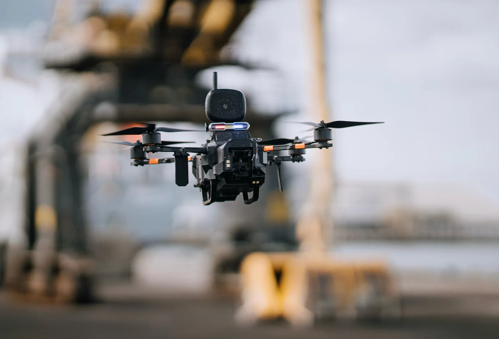 Black tactical quadcopter with a spherical sensor pod and red-and-blue strobe lights hovering in front of an industrial port crane