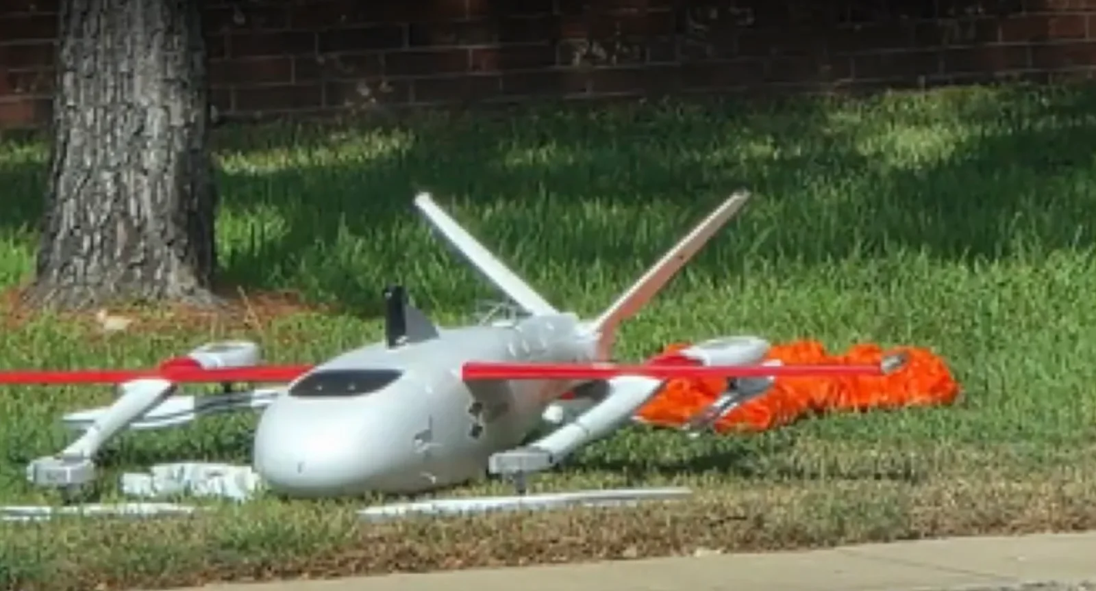 White Zipline Zip delivery drone resting on grass with red wing spar, rotor booms and tail intact, and an orange parachute bundled beside the fuselage