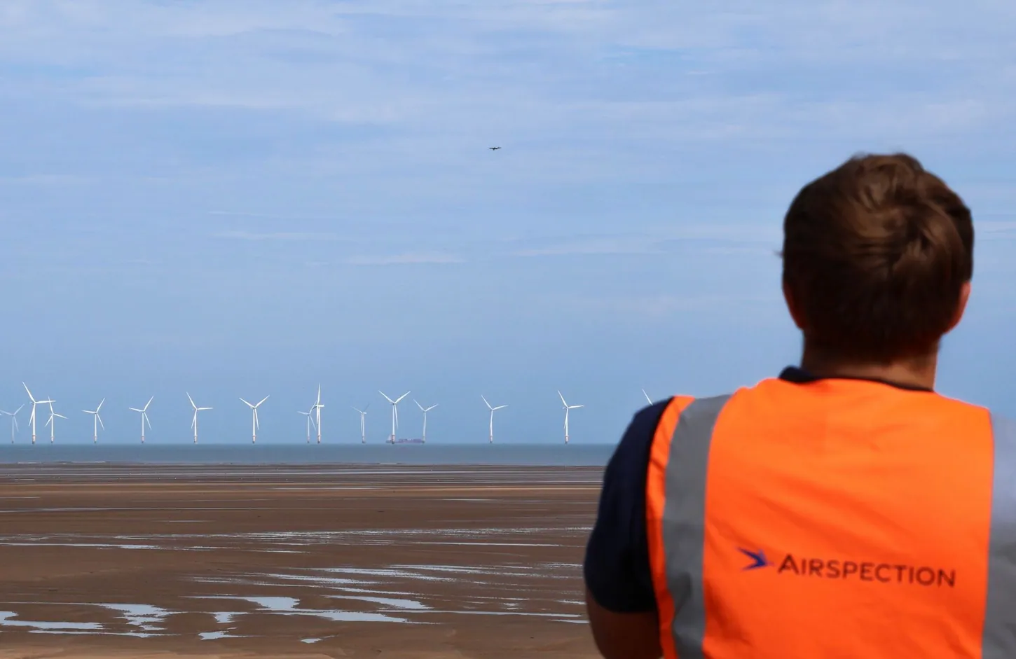 Airspection crew member in an orange high-visibility vest on a tidal beach watching a small drone in the sky, with offshore wind turbines on the horizon