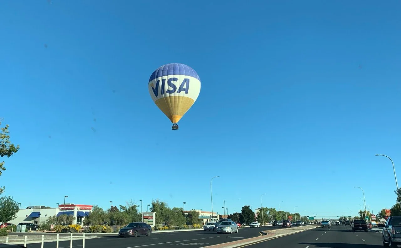 A blue, white and gold VISA hot air balloon floats in a clear blue sky above a multi-lane road and roadside businesses in Albuquerque.