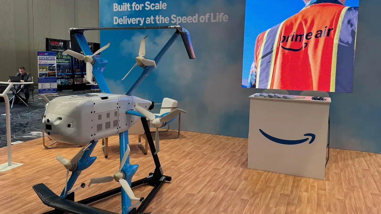 Amazon Prime Air MK30 delivery drone on a black display stand at a trade show booth, with a Prime Air vest on a screen behind it