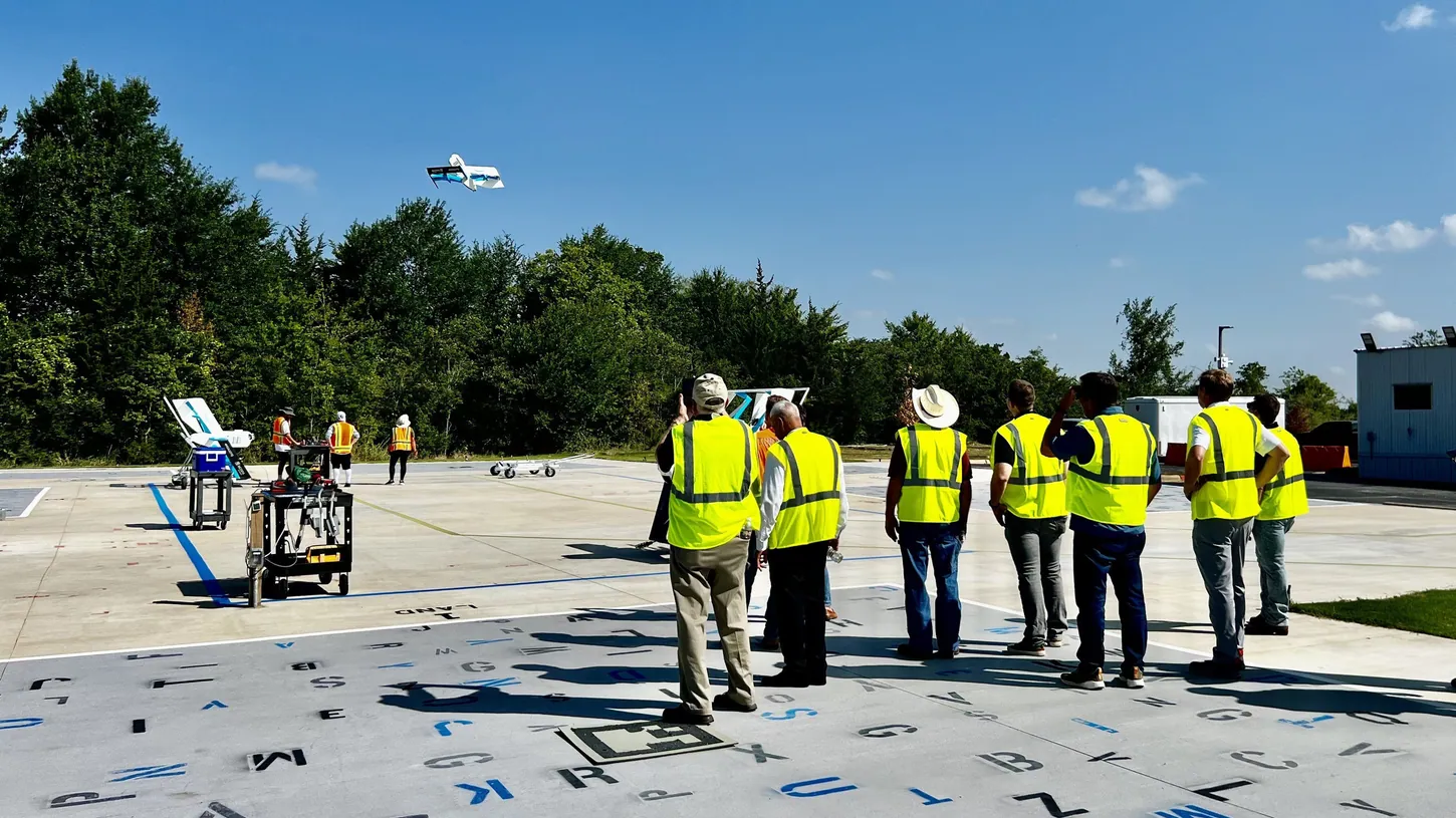 A group of people in yellow safety vests on a concrete test pad watch a white and blue Amazon MK30 delivery drone fly above a tree line