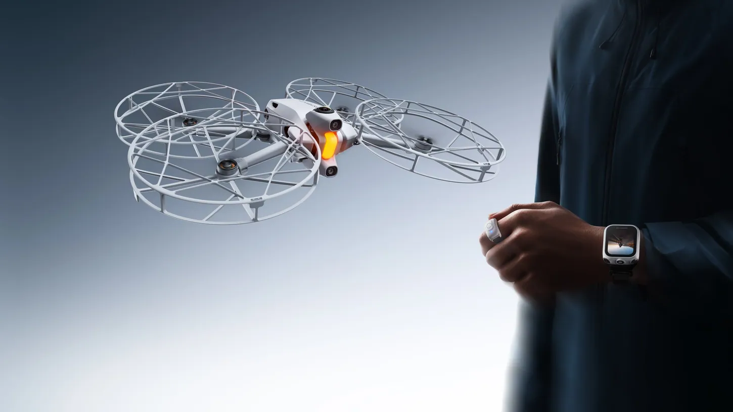 White Antigravity A1 drone with four full-coverage propeller guards hovering next to a person in a dark jacket wearing a watch remote and a ring remote