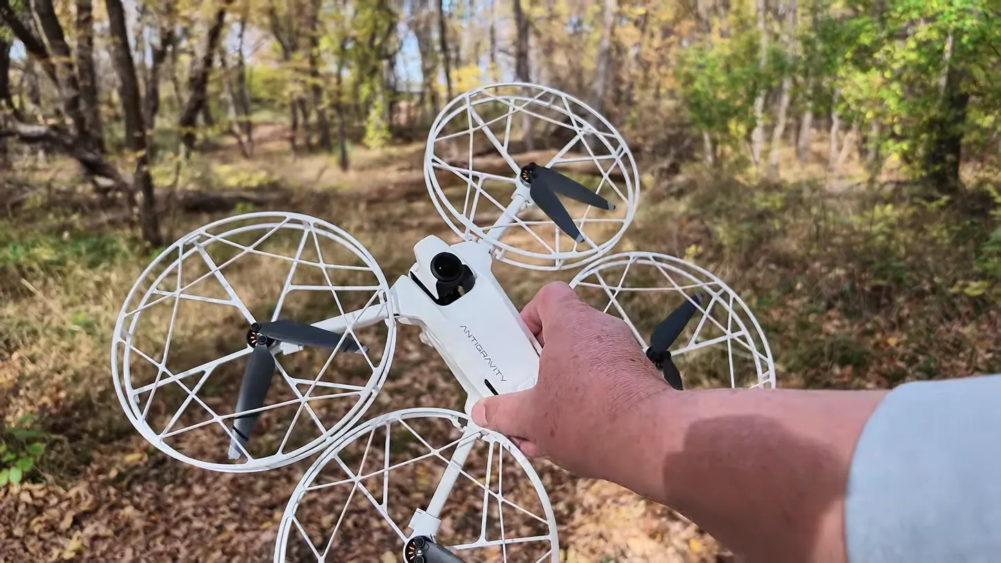 Hand holding a white Antigravity A1 drone fitted with four white cage-style propeller guards on a leaf-covered wooded trail