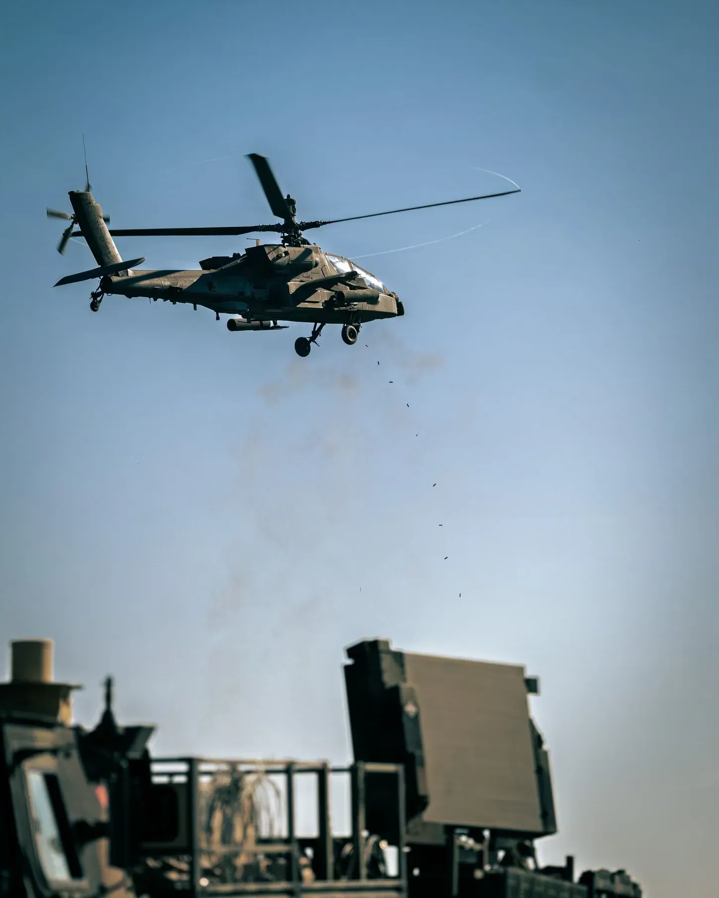 AH-64 Apache helicopter firing its chin-mounted 30mm cannon, spent casings falling below it, with the flat panel of a Sentinel radar on a vehicle in the foreground