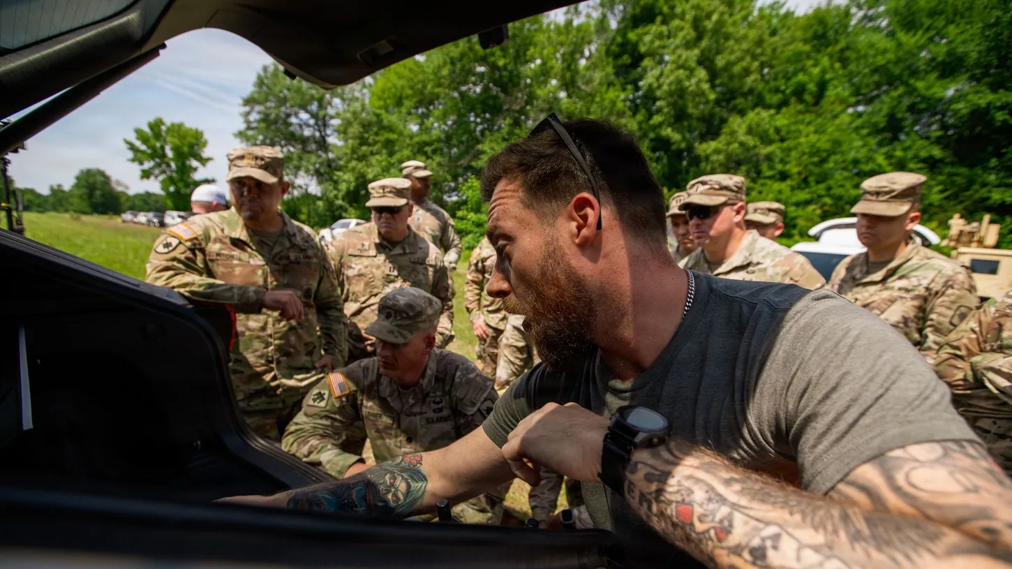 A bearded instructor in a gray T-shirt leans into the open trunk of a vehicle holding counter-drone equipment while a group of Army National Guard soldiers in camouflage uniforms watches behind him in a grassy field