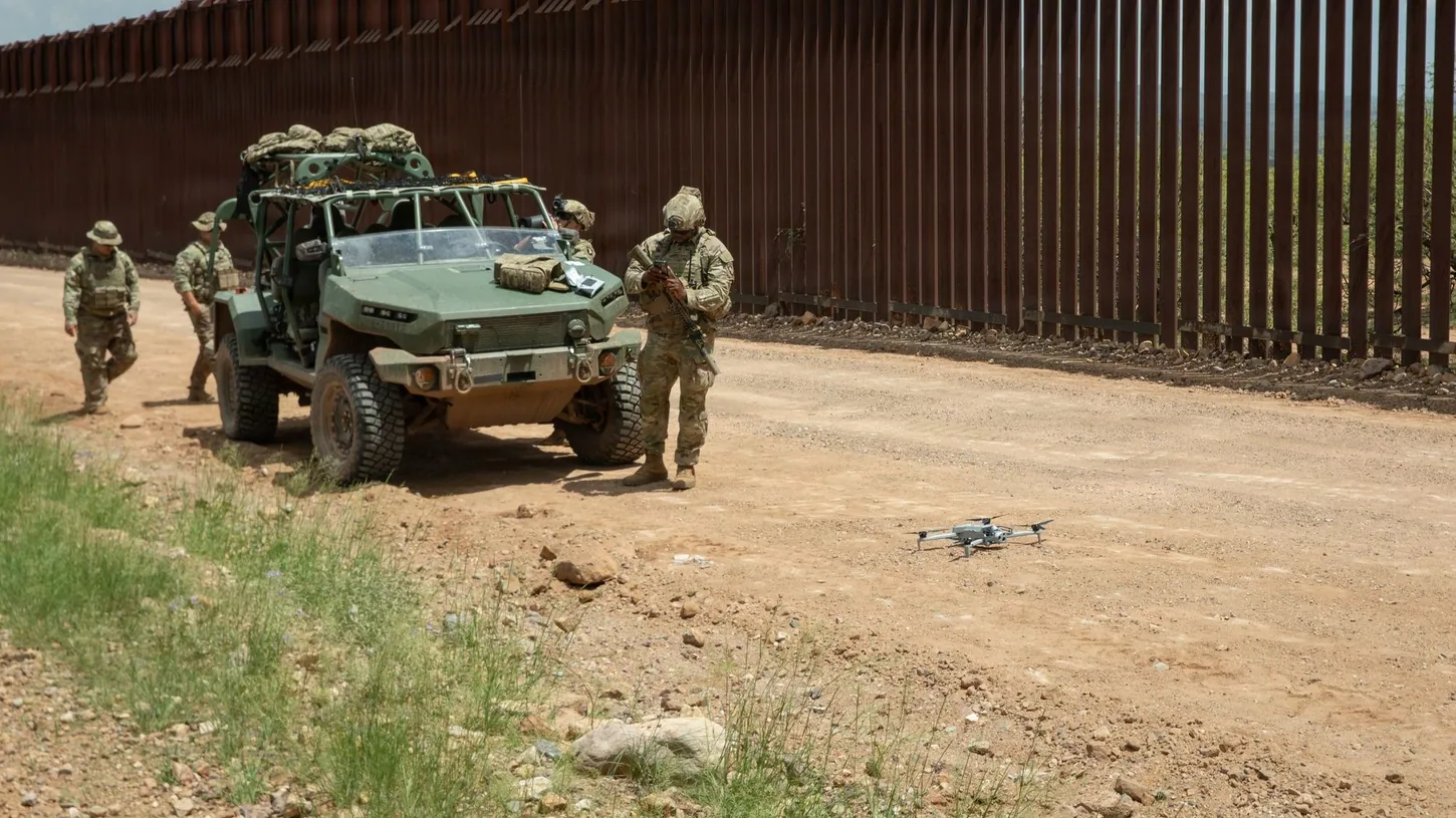 Soldiers in camouflage stand beside a green military utility vehicle on a dirt road along a tall steel border wall, with a gray quadcopter drone resting on the ground in the foreground.