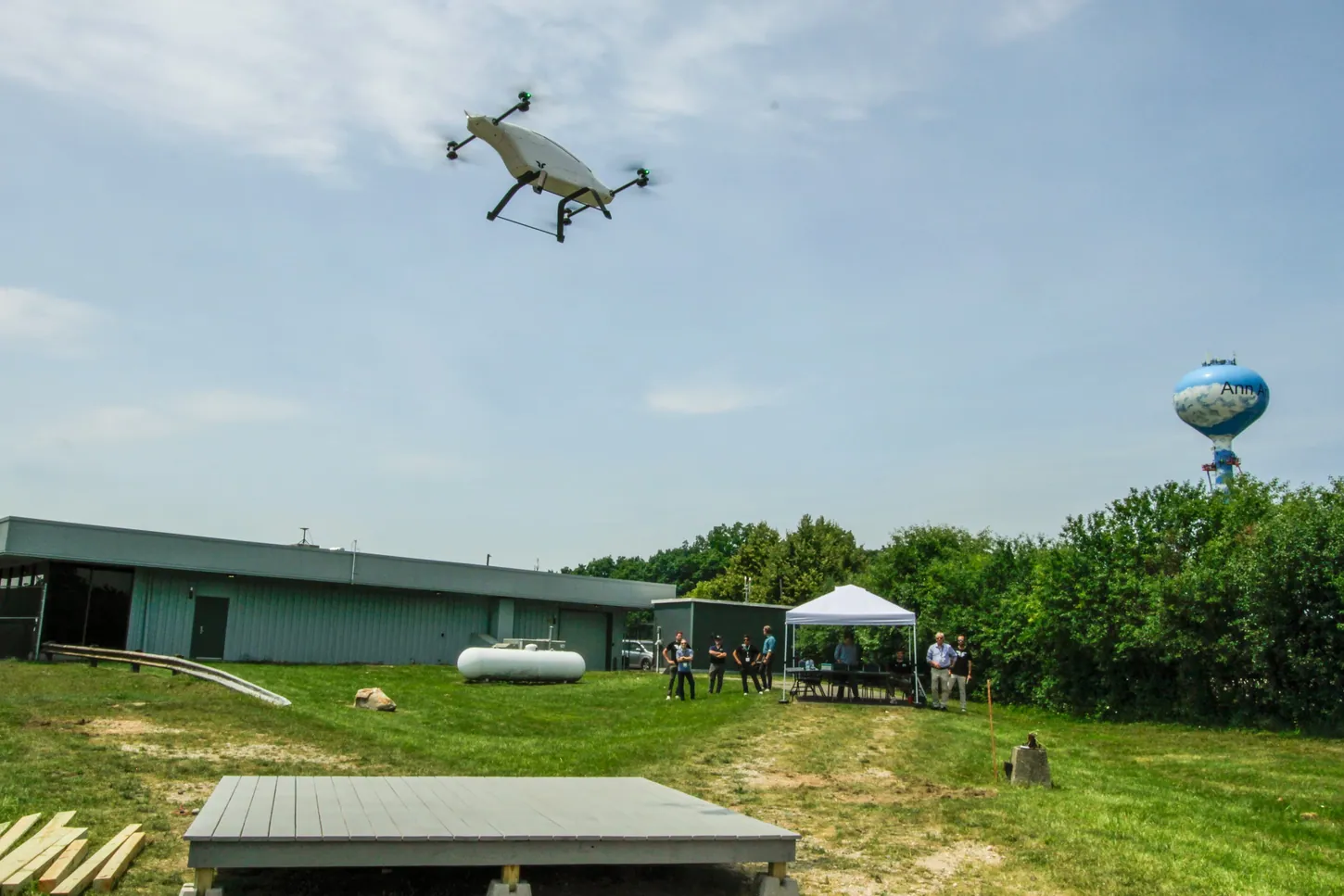 White blueflite drone flying over a grass field at the M-air proving grounds as a group of people watches from beside a white tent
