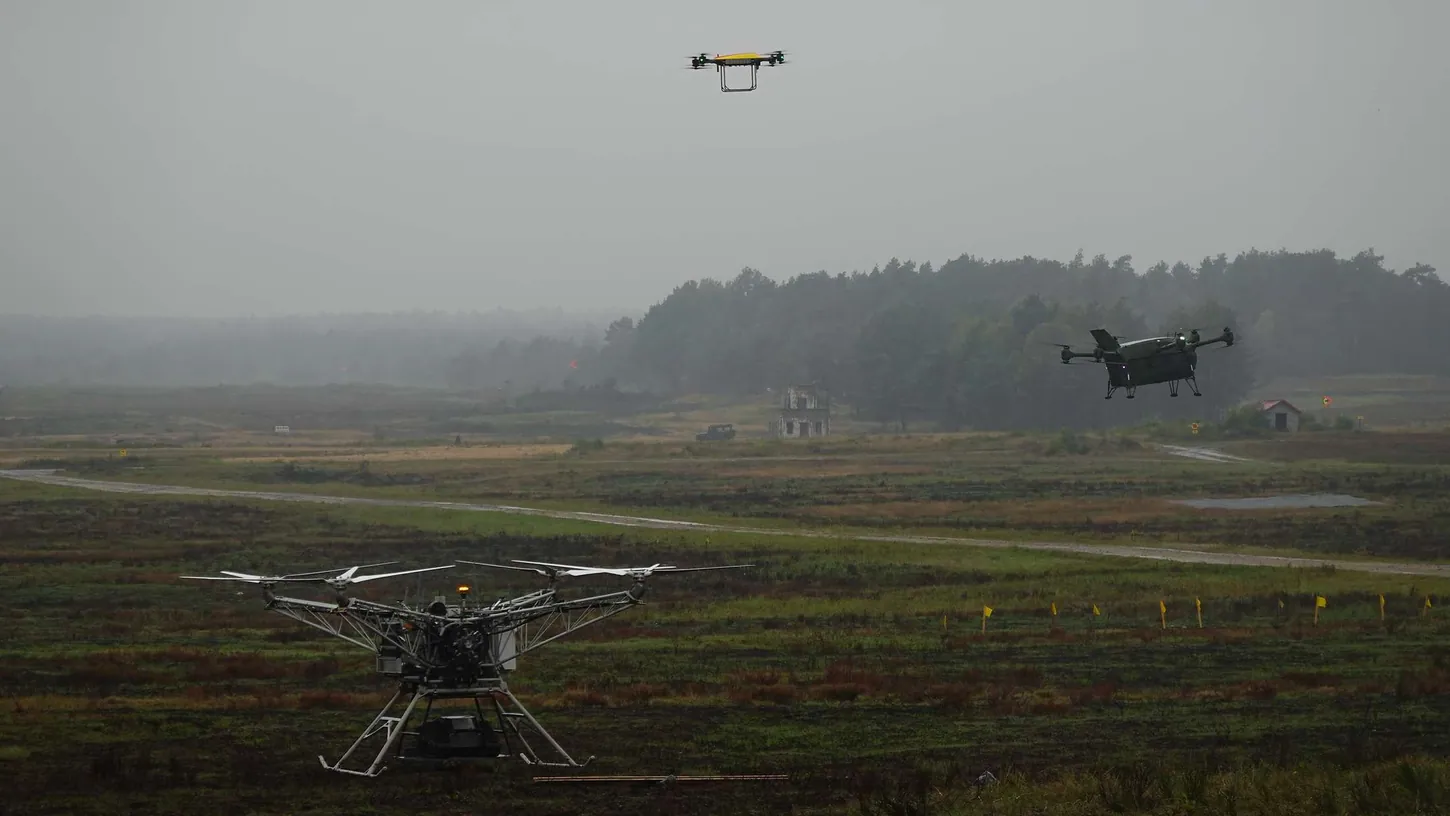 Three drones of different sizes, including a large multirotor on the ground and two quadcopters in the air, over a rainy military training area