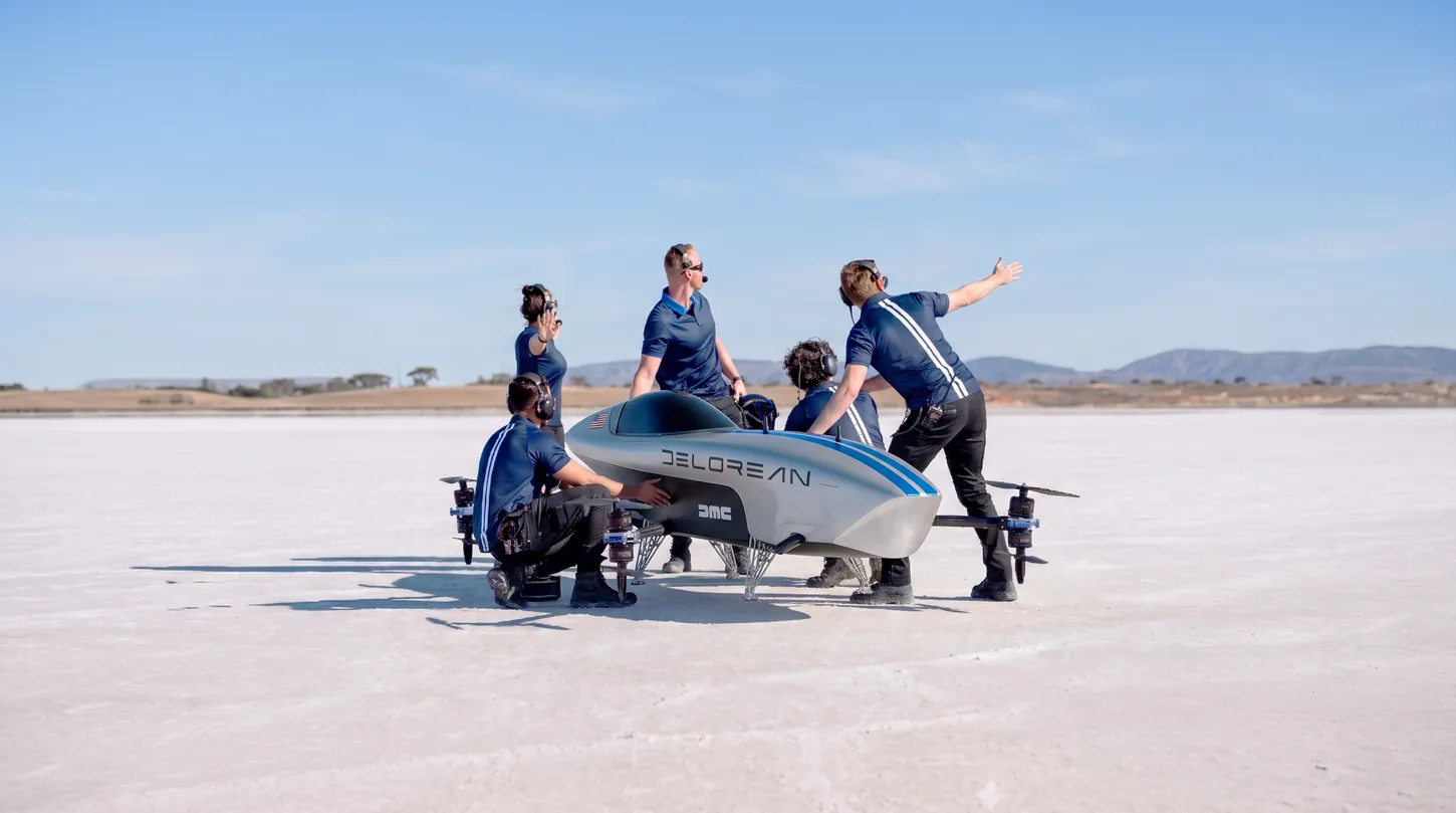 Five crew members in navy team shirts and headsets working around the DeLorean-liveried Airspeeder Mk3C on a salt lake