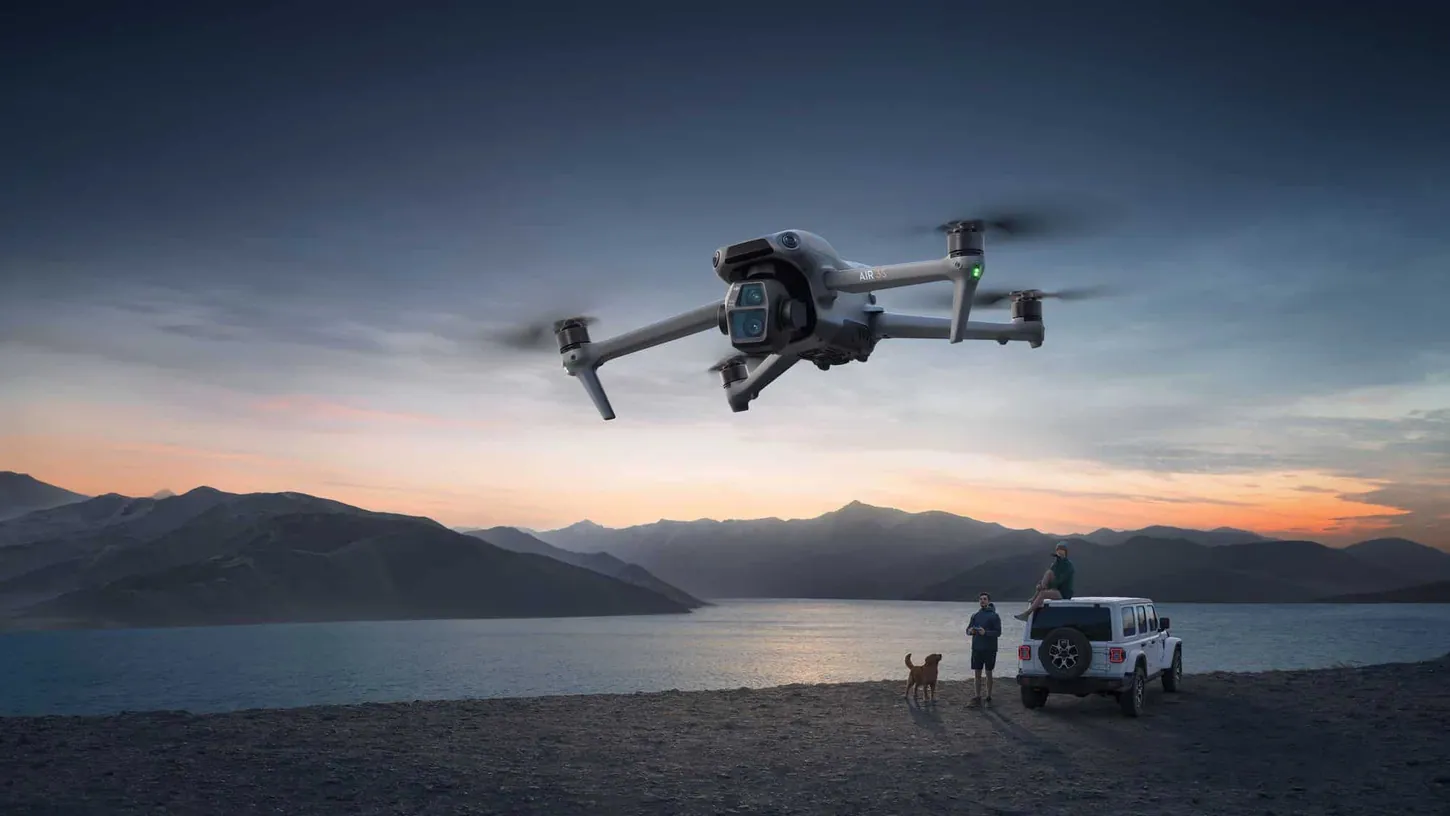 DJI Air 3S drone flying over a lakeshore at dusk while a pilot holding a controller, a dog and a white SUV wait below