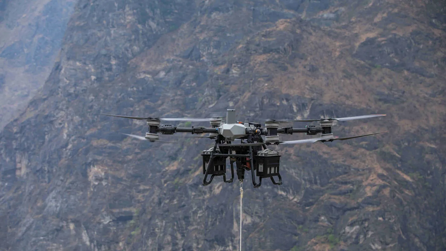 Gray eight-rotor DJI FlyCart 100 cargo drone hovering in front of a rocky mountainside with a winch cable hanging below it