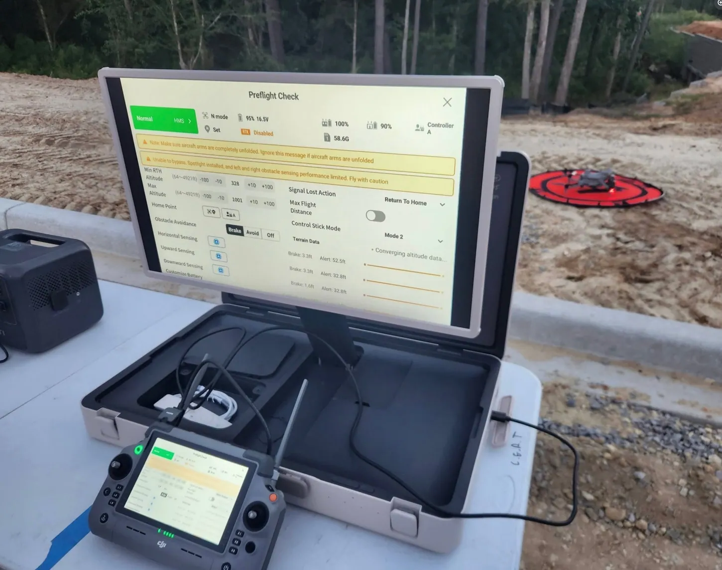 DJI Pilot 2 preflight check screen on a large monitor in an open case, with a DJI RC Plus 2 controller in front and a Matrice 4T on a red landing pad in the dirt behind