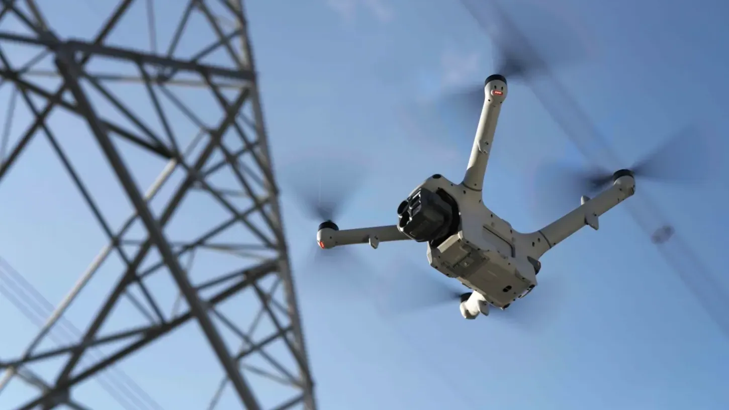 DJI Matrice 5TD drone seen from below in flight beside a steel transmission tower against a blue sky