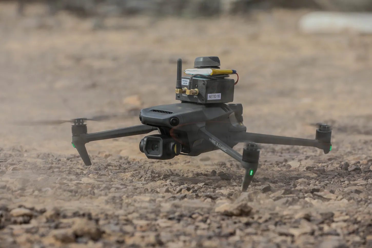 A gray DJI Mavic 3 Classic with a small radio module strapped on top hovers low over gravel desert ground.