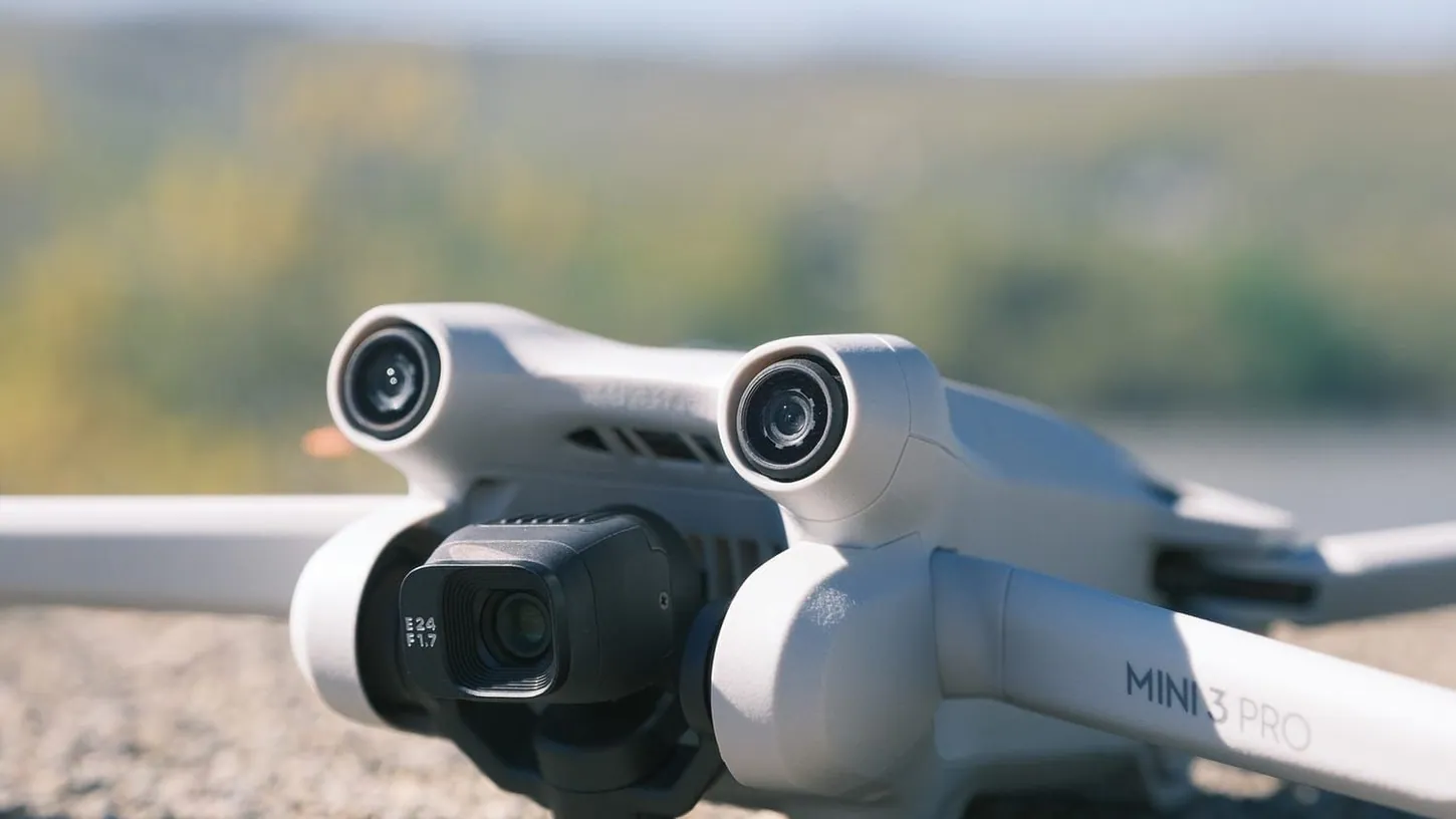 Close-up of a white DJI Mini 3 Pro on a stone ledge, showing its gimbal camera, twin forward obstacle sensors and the Mini 3 Pro label on one arm, with a blurred river valley behind.