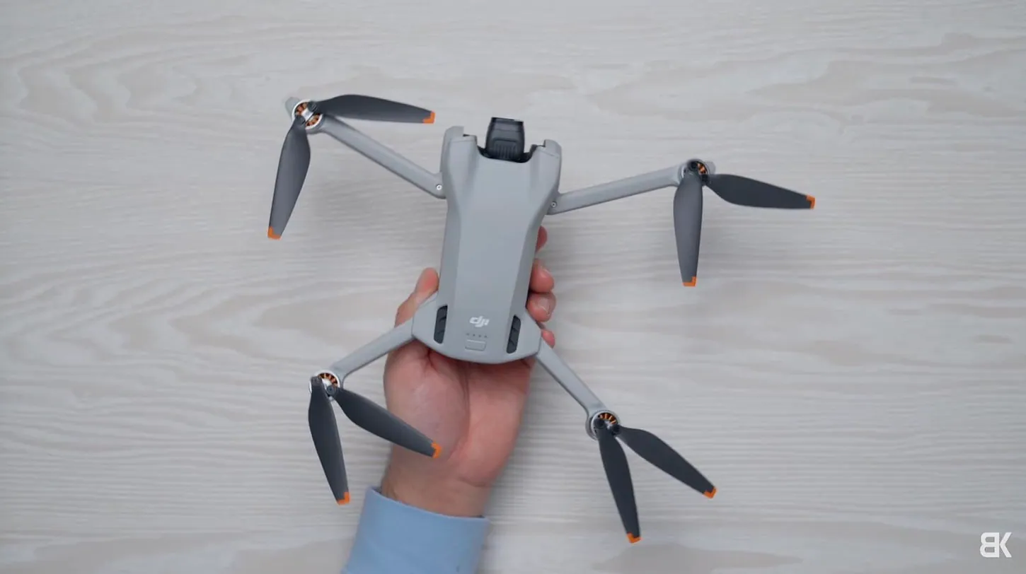 DJI Mini 3 seen from above with its arms and propellers unfolded, held in one hand over a white wooden table