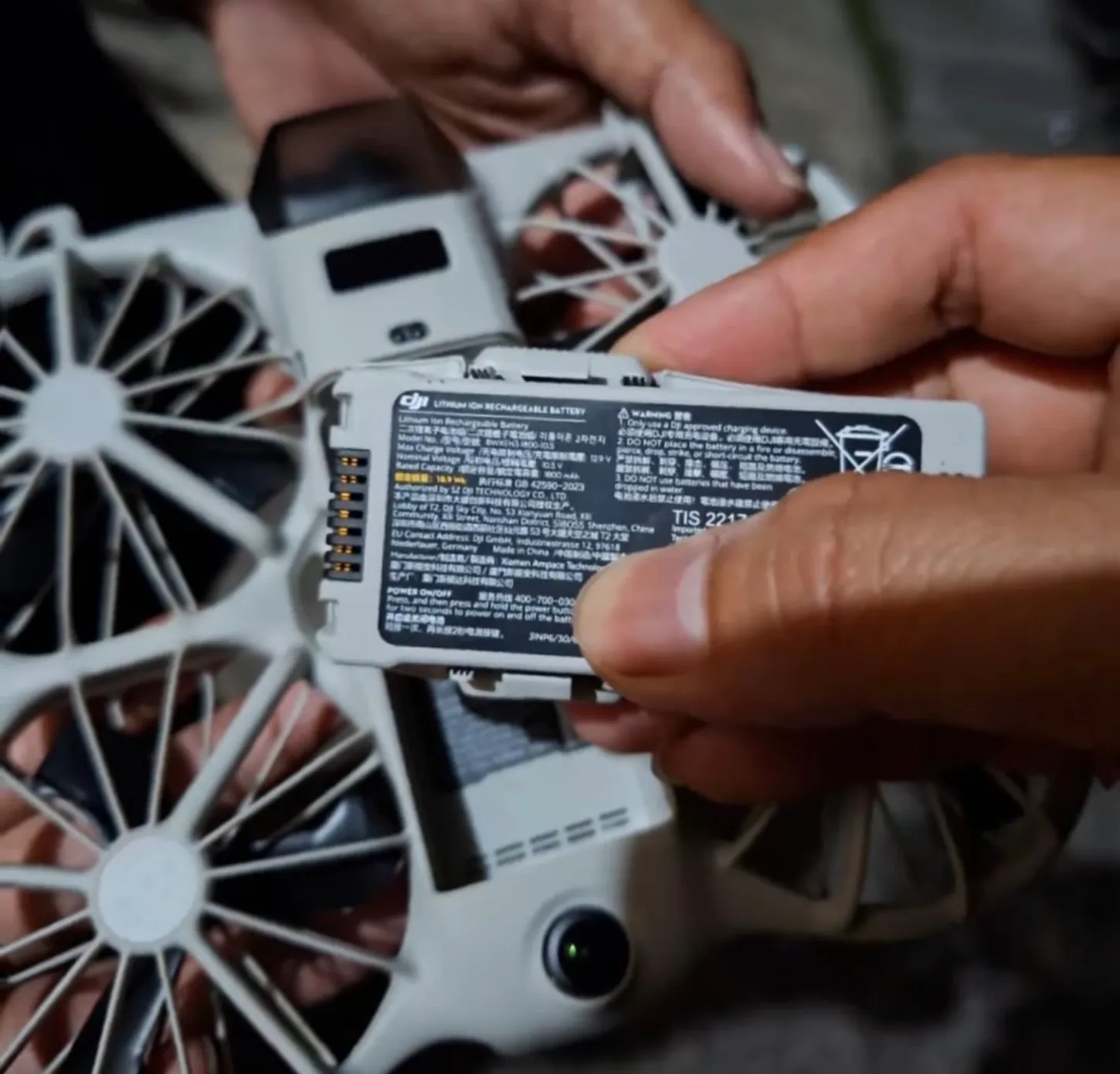 Hand holding a DJI Neo 3 flight battery with its black label facing the camera, showing a 1,800 mAh rating, 18.9 Wh, a row of gold contacts on one end and the caged drone behind it