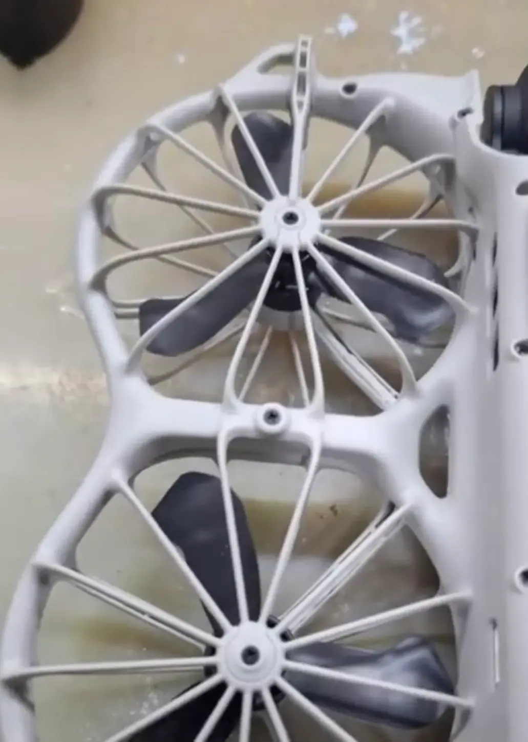 Underside of two white DJI Neo 3 propeller cages on a dusty tabletop, with a screw in the centre of each hub, a screw where the cages join and black propeller blades inside the spokes