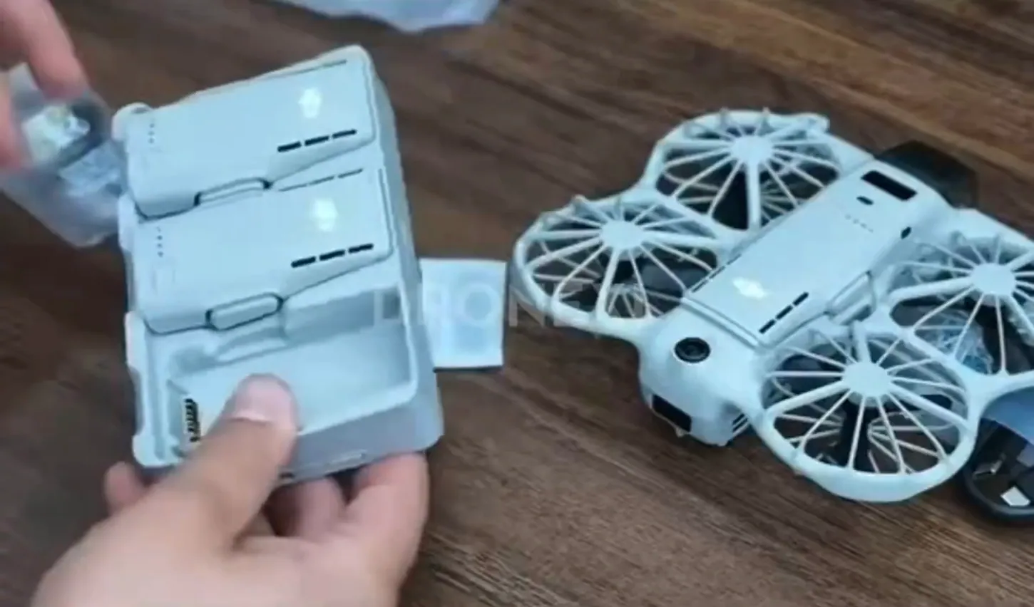Hand holding the DJI Neo 3 charging hub with two flight batteries next to the caged Neo 3 drone on a wooden table