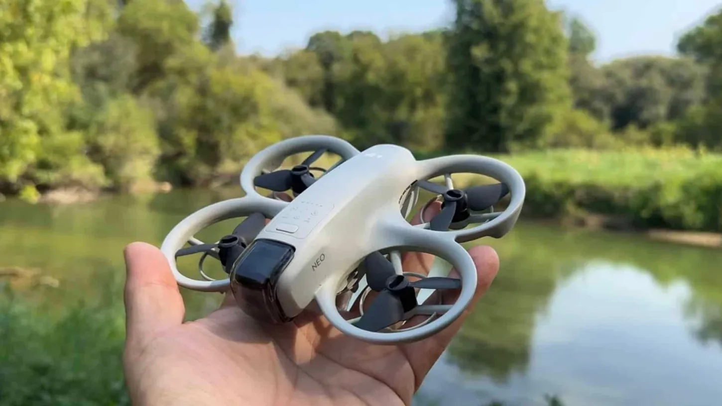 DJI Neo drone resting on an open palm with a river and trees behind it